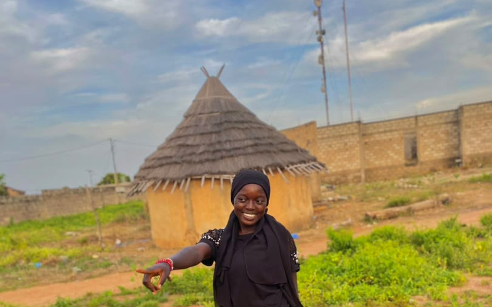 Rima smiling in front of a thatched-roof hut in her village